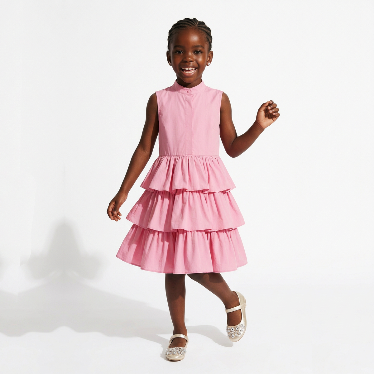 A young girl smiling and posing in a sleeveless pink tiered dress with a high mock neck, paired with sparkly silver shoes, against a clean white studio background.