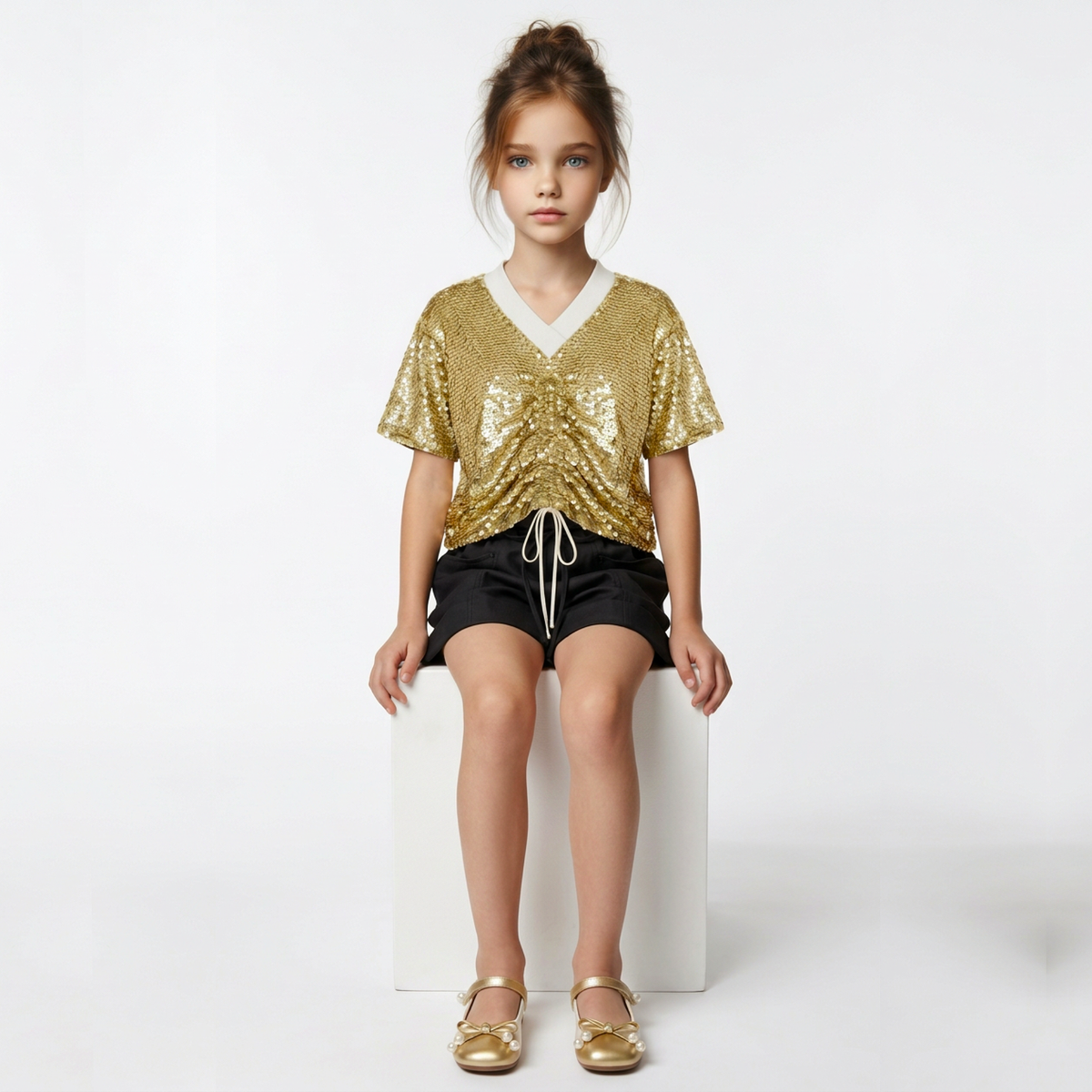 A young girl sits on a white block against a white studio background, wearing a gold sequined short-sleeve top with a white V-neck trim, black shorts, and gold flat sandals.
