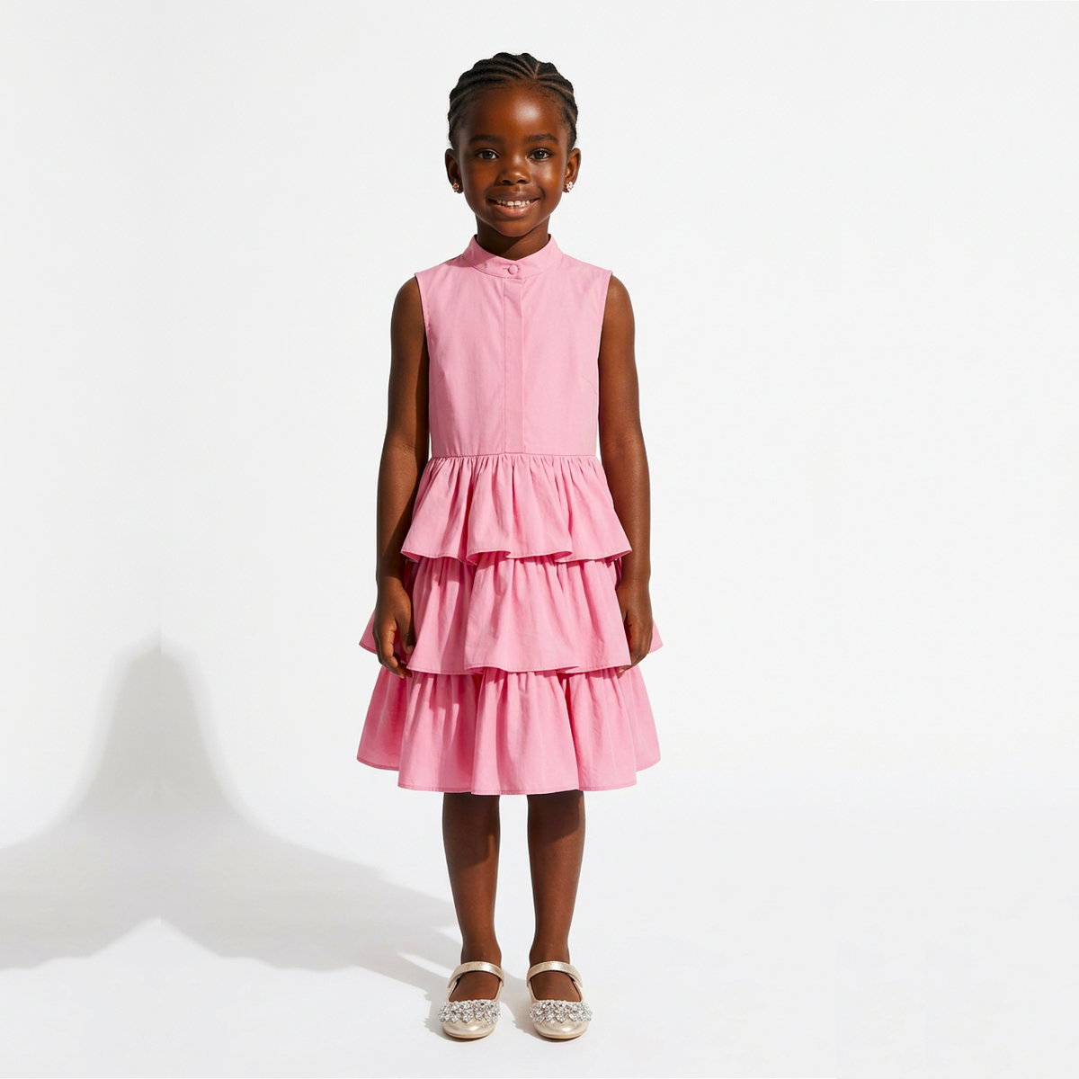 A young girl with braided hair stands against a white studio background wearing a sleeveless pink tiered dress with a high mock neck and light-colored flat shoes.