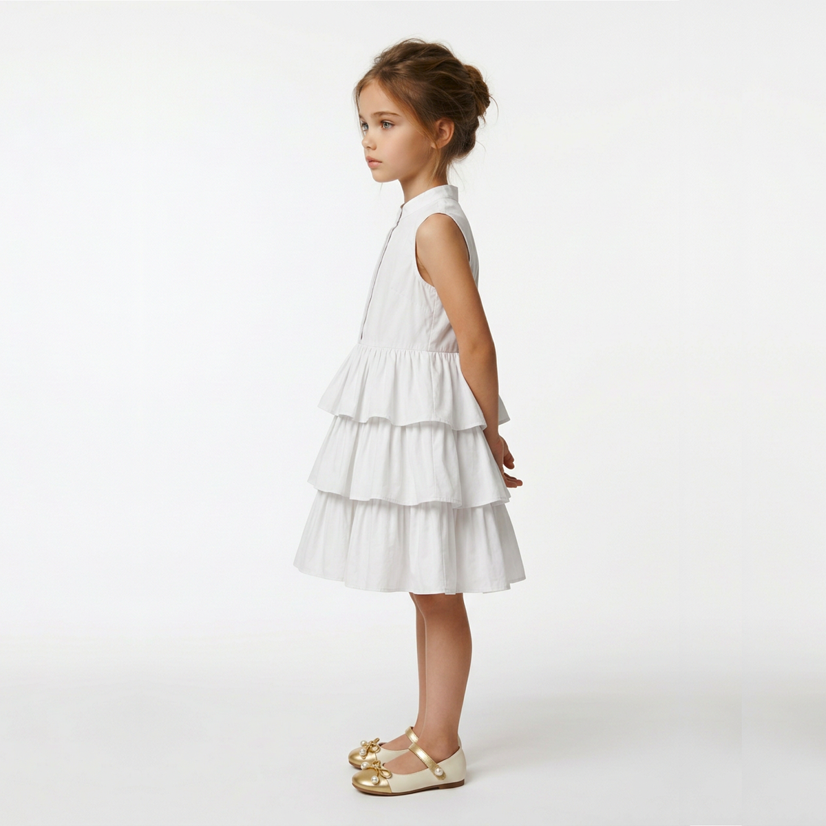 A young girl with her hair in a bun is shown in profile wearing a sleeveless white tiered dress and gold Mary Jane shoes, standing against a seamless white studio background.