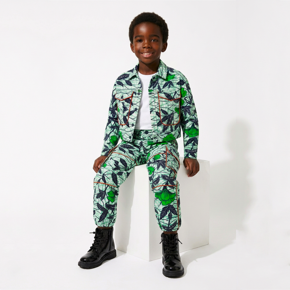 A young boy wearing a matching green tropical leaf print jacket and pants set with black boots, sitting on a white block against a white studio background.