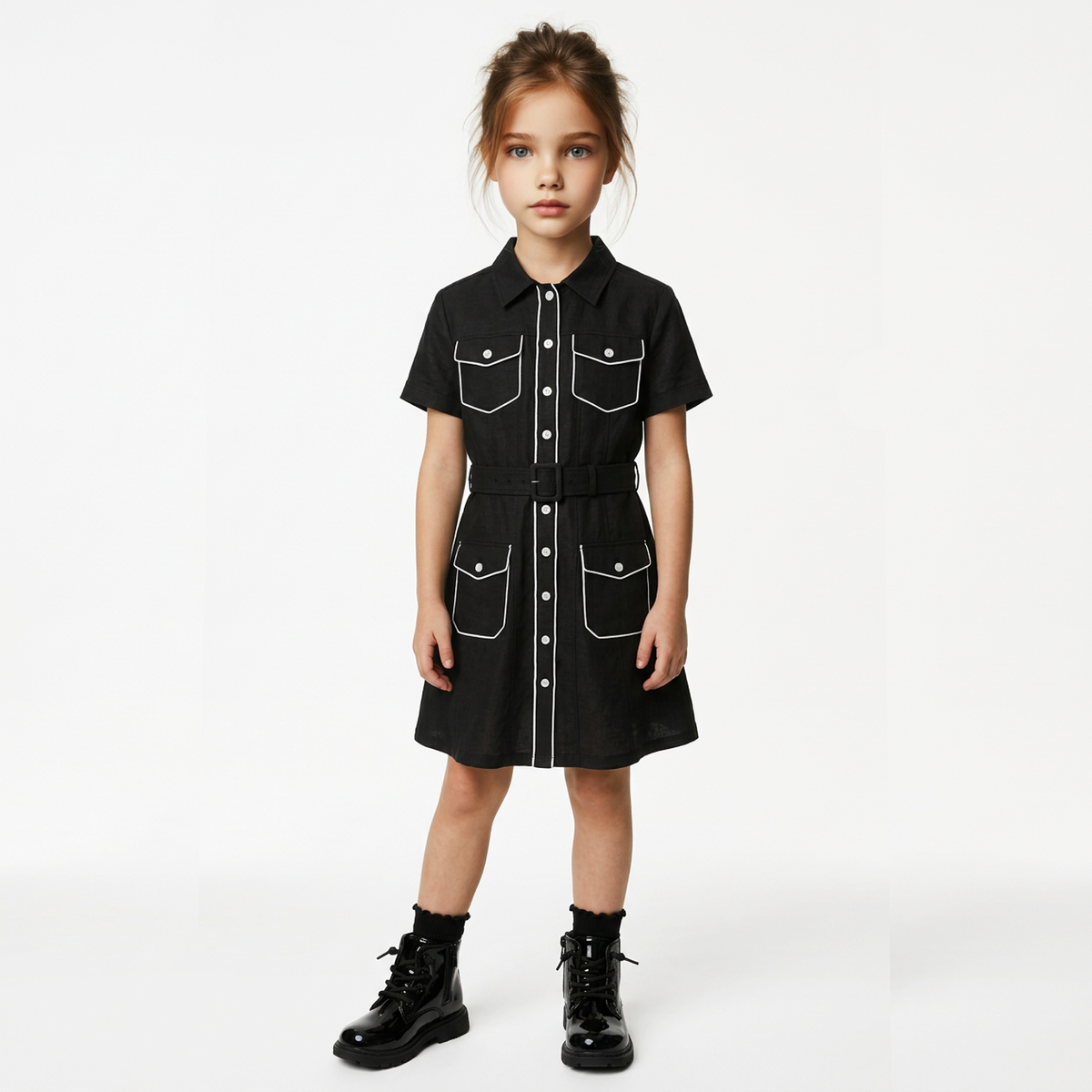 A young girl stands against a plain white studio background wearing a black short-sleeve button-front dress with white contrast stitching, flap chest pockets, side pockets, a matching black belt, and black lace-up boots.