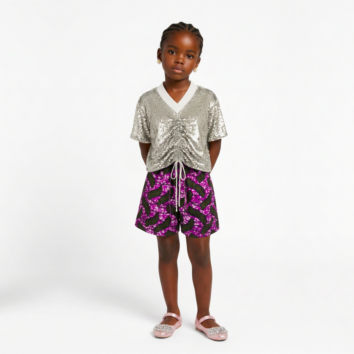 A young girl stands against a light gray studio background wearing a silver sequined short-sleeve top with a white V-neck and purple-and-black patterned shorts.