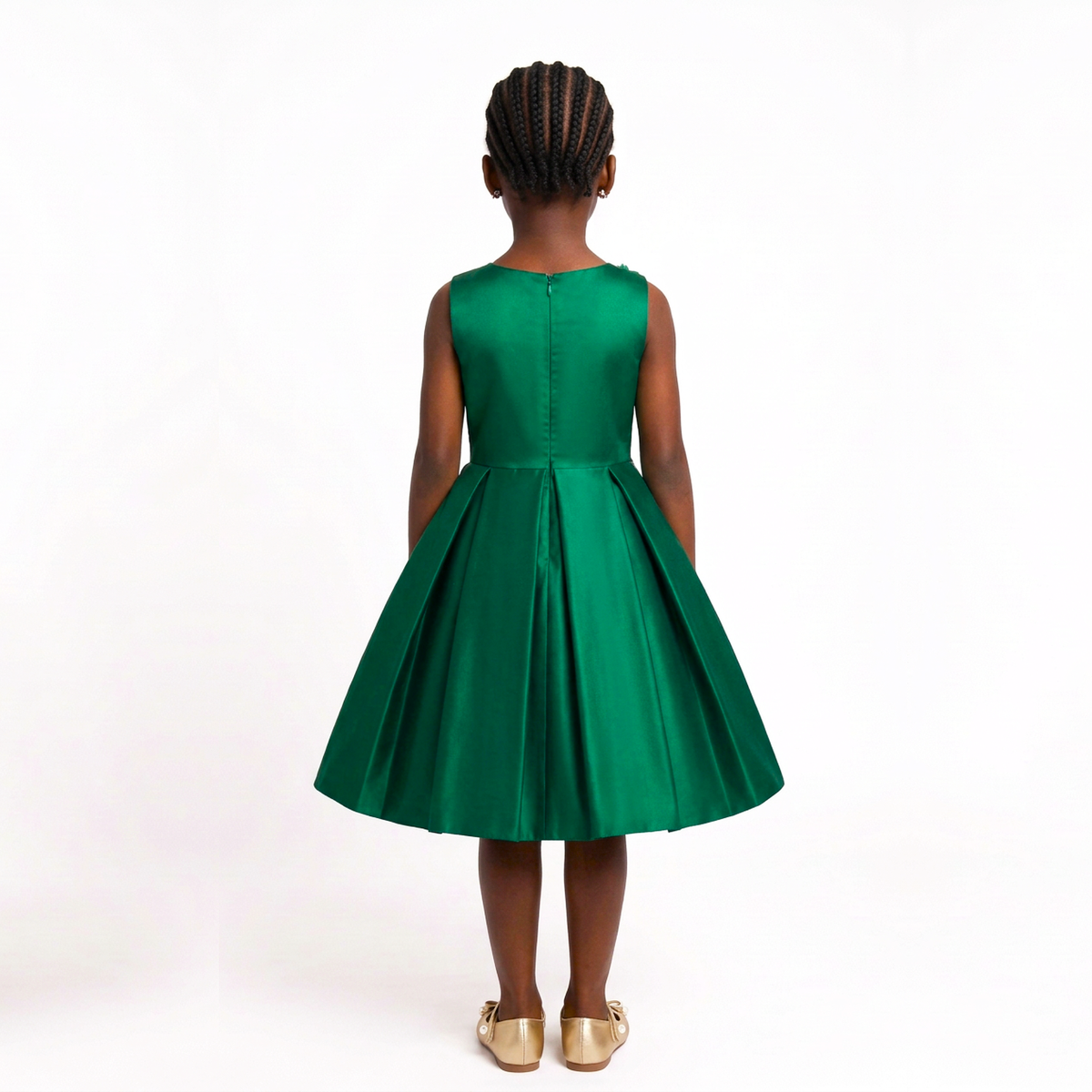 A young girl viewed from behind wearing a sleeveless emerald green dress with a pleated skirt, paired with gold shoes, against a plain white studio background.