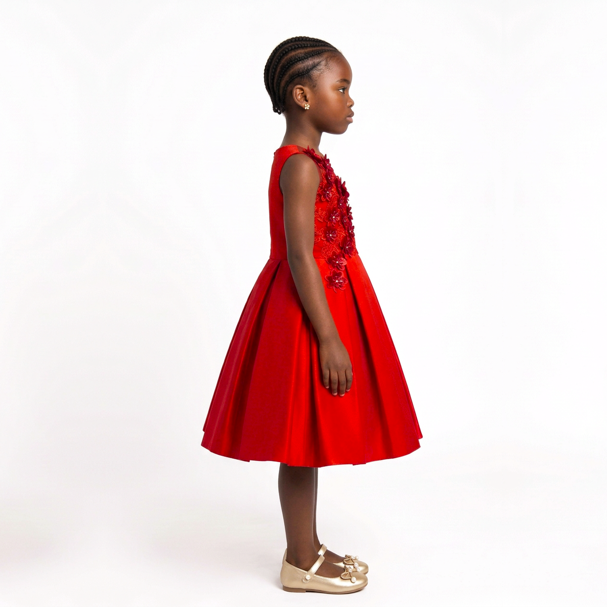 A young girl in profile wearing a bright red sleeveless dress with 3D floral applique on the bodice, paired with beige Mary Jane shoes, photographed against a seamless white studio background.