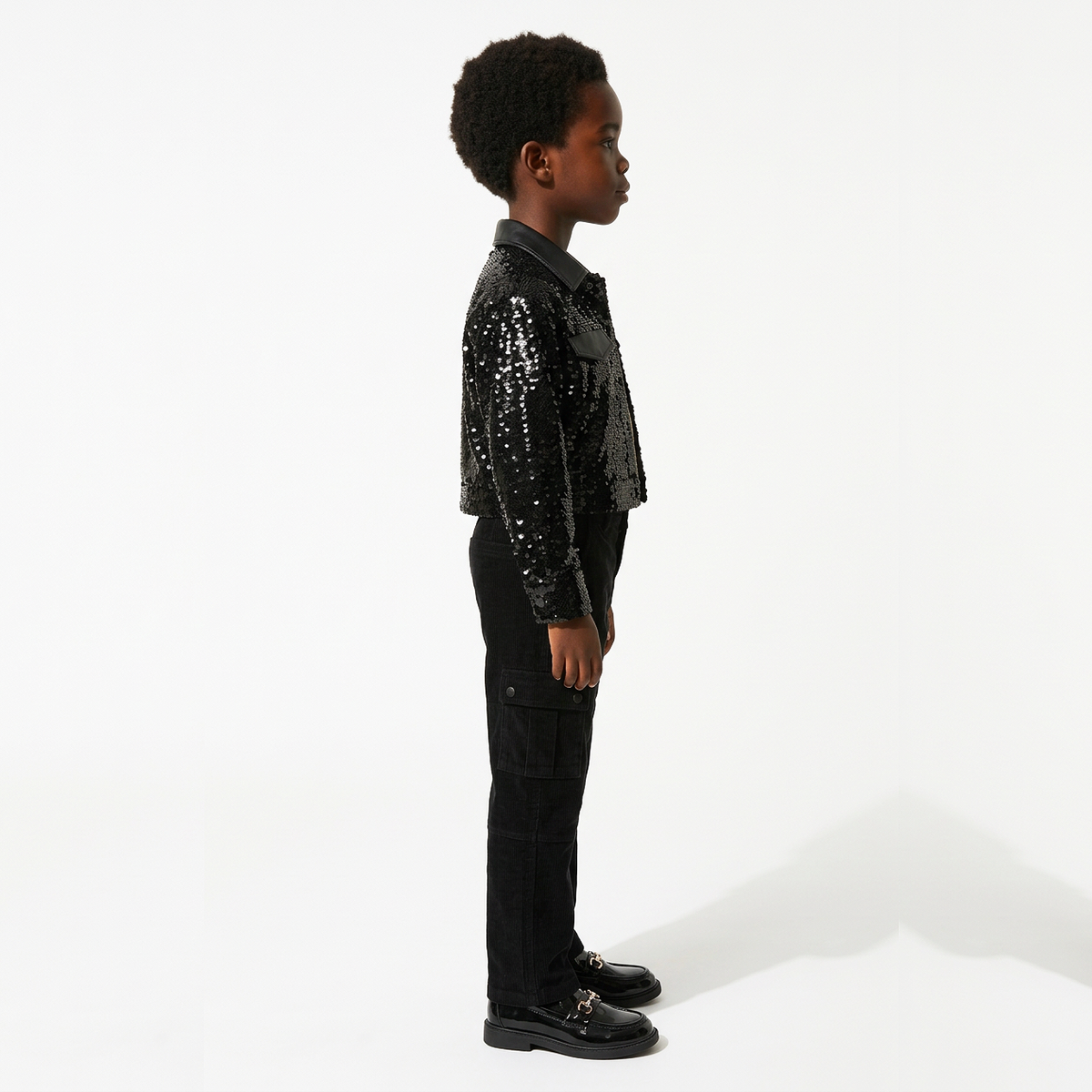A child model shown in profile wearing a black sequined jacket with a dark collar, black cargo pants, and black shoes, standing against a clean white studio background.