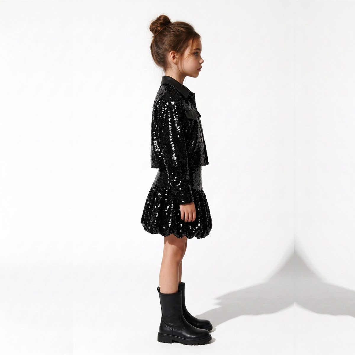 A young girl with her hair in a bun stands in profile wearing a black sequined jacket and matching sequined mini skirt, paired with black ankle boots, against a clean white studio background.