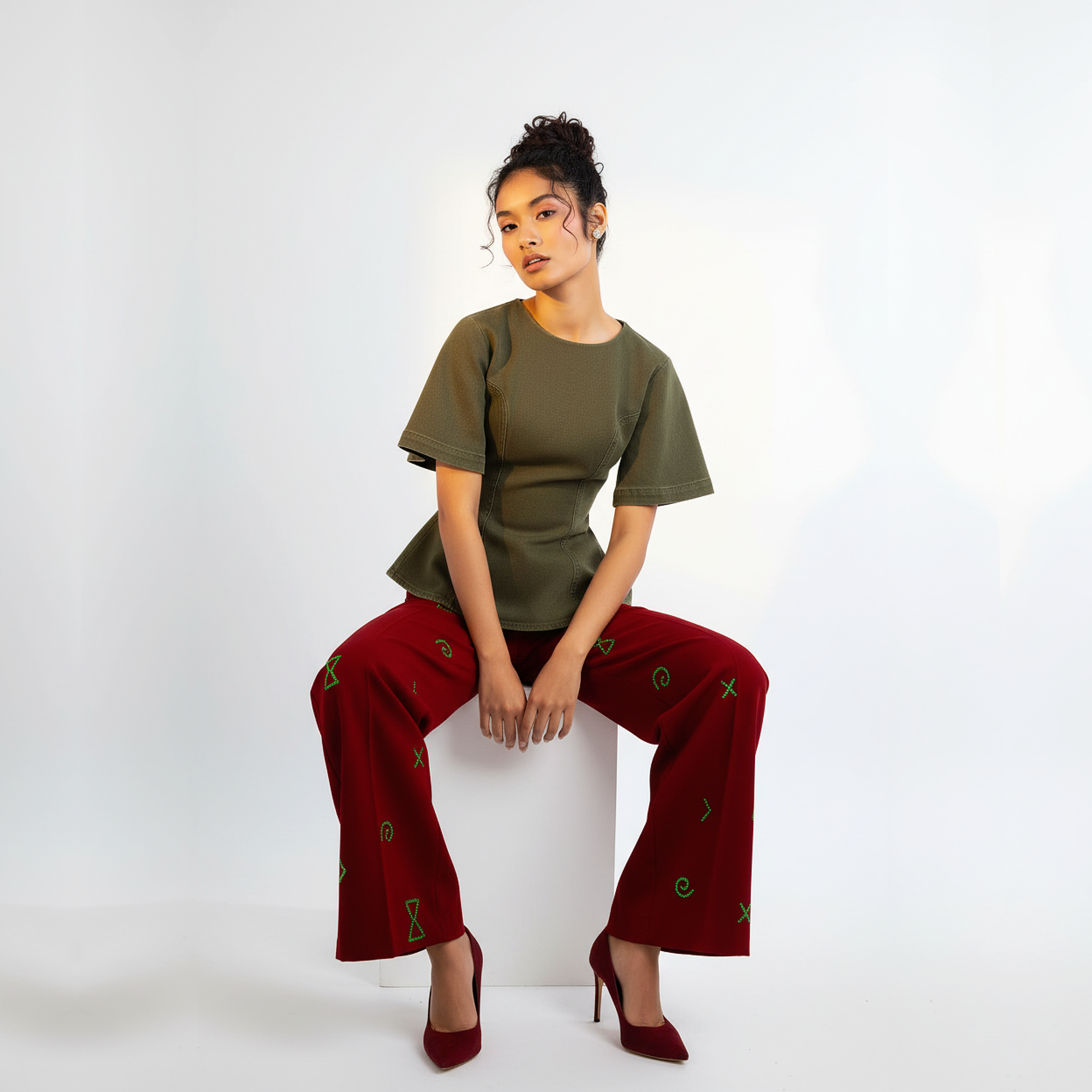 Woman wearing a green top and red pants sitting on a white stool against a white background