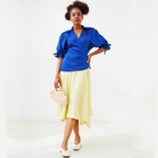 Woman wearing a dress with a  blue top and yellow skirt holding a beige handbag on a white background