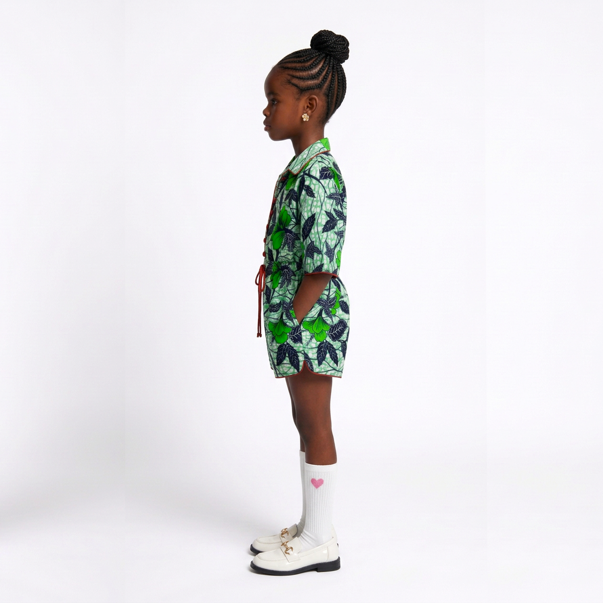 A young girl in profile wearing a green and white tropical leaf print short-sleeve collared shirt and matching shorts, styled with white socks and white shoes, against a plain white studio background.