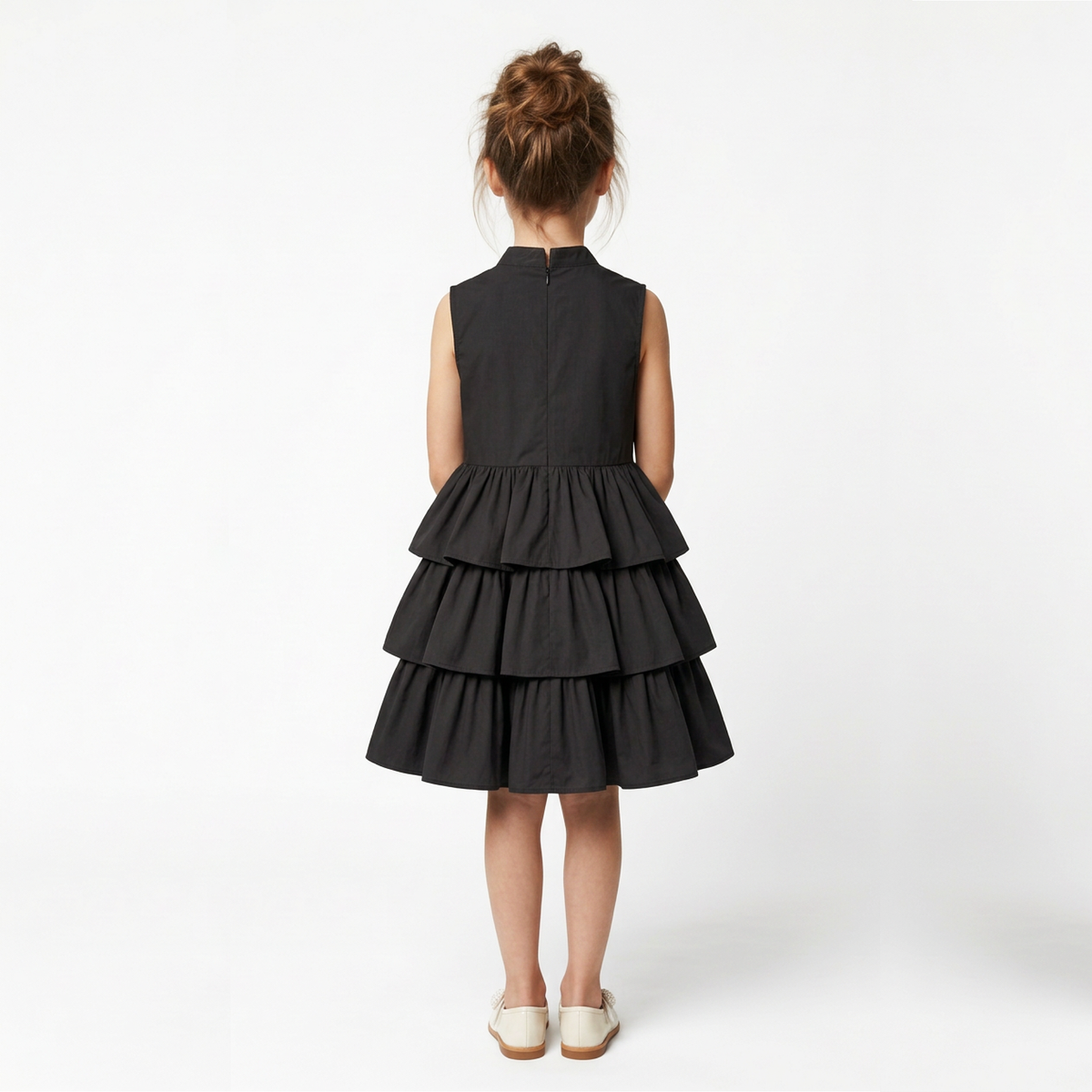 A young girl viewed from behind wearing a sleeveless black dress with a tiered ruffle skirt, paired with off-white shoes, against a plain white studio background.
