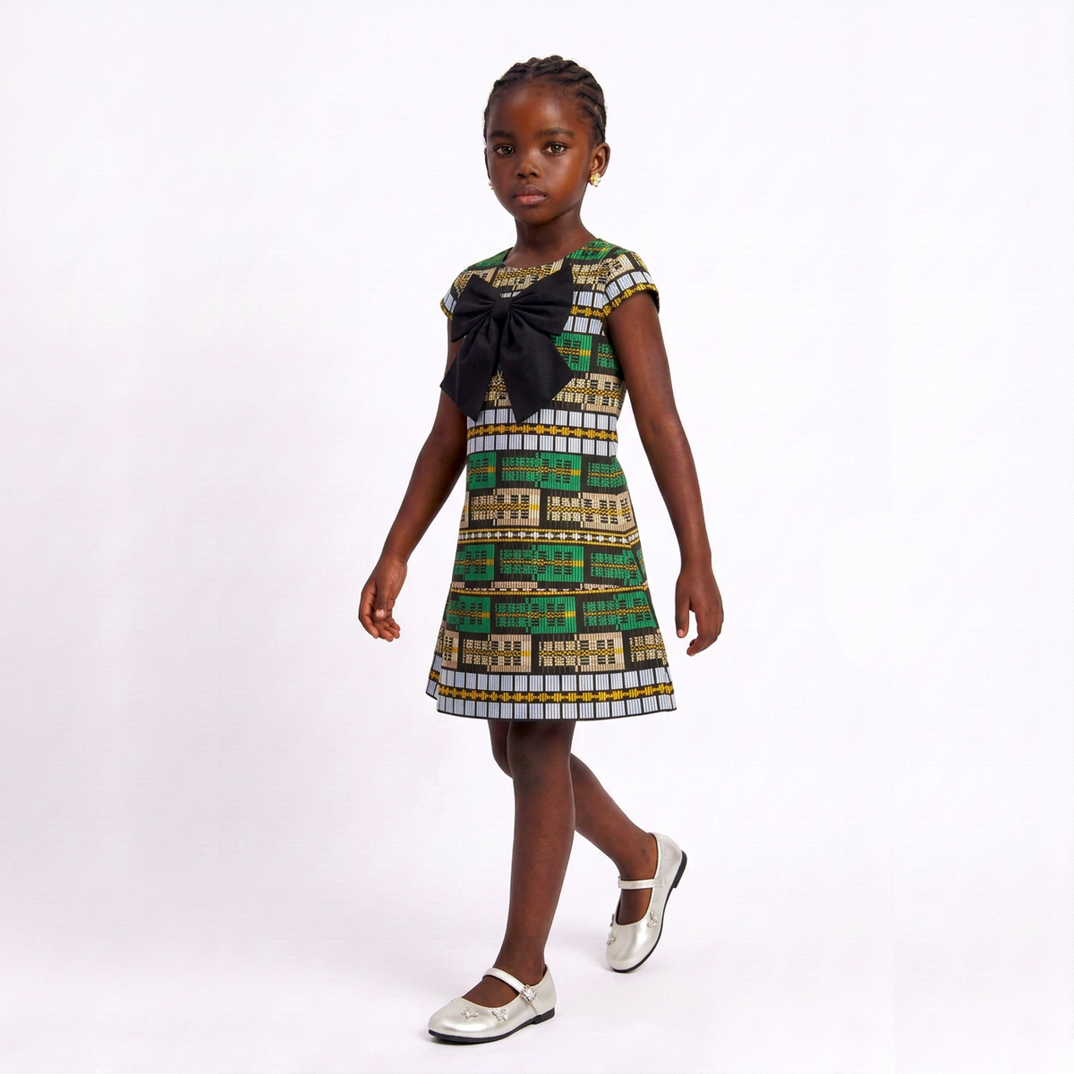 A young girl modeling a sleeveless dress with a colorful African woven Akwete print pattern featuring green, gold, and white geometric motifs, accented with a large black bow at the neckline. She is wearing white Mary Jane shoes against a plain white studio background.