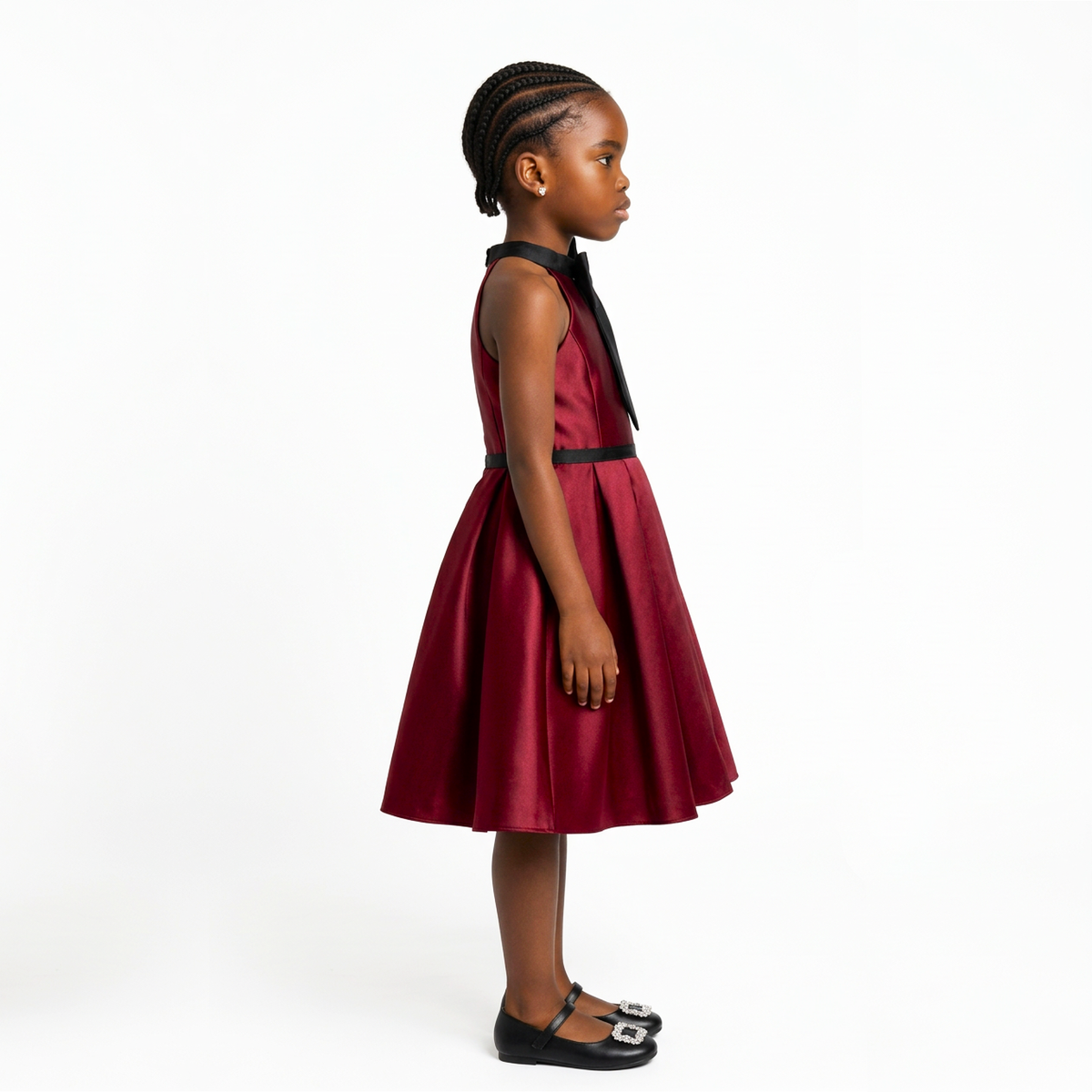 A young girl with braided hair stands in profile wearing a sleeveless, pleated dark red dress with a black collar ribbon and a thin black waistband, paired with black Mary Jane shoes on a white studio background.