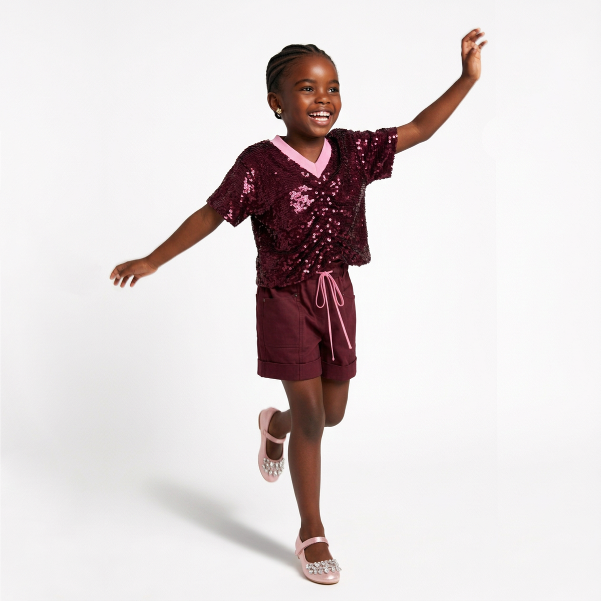 A young girl smiling and posing in a sparkling burgundy sequined short-sleeve crop top and matching shorts set, styled with a light pink collar, matching pink drawstring, and pink glittery Mary Jane shoes against a white studio background.