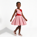 A young girl modeling a sleeveless pink dress with a red diagonal bodice panel and a matching pink bow on the back, paired with pink sparkly flats, against a clean white studio background.