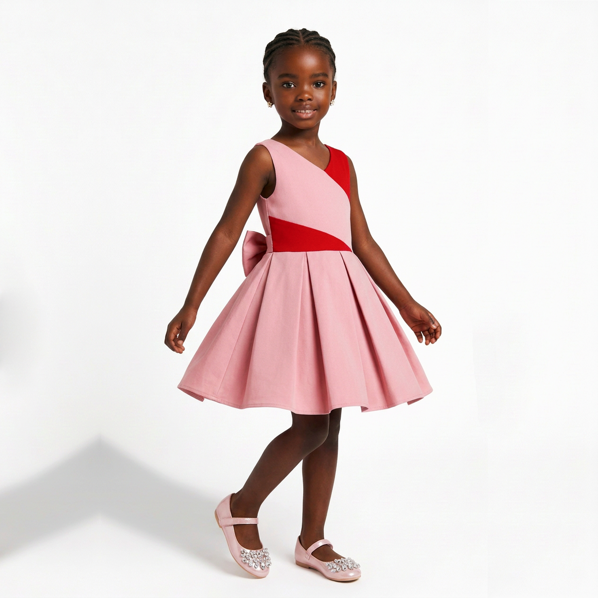 A young girl modeling a sleeveless pink dress with a red diagonal bodice panel and a matching pink bow on the back, paired with pink sparkly flats, against a clean white studio background.