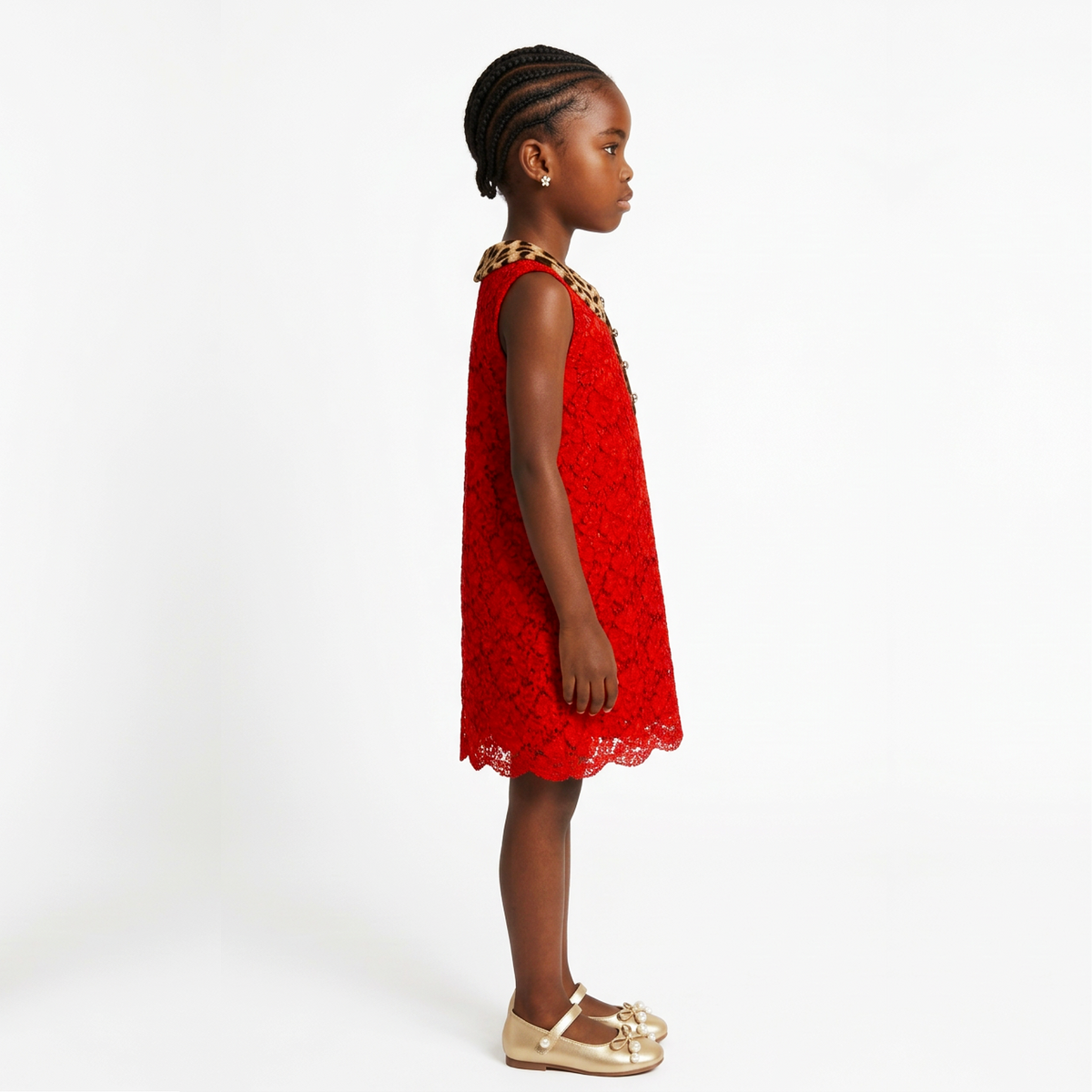 A young girl in profile wearing a sleeveless red lace dress with a gold chain collar and gold flat shoes, photographed against a clean white studio background.