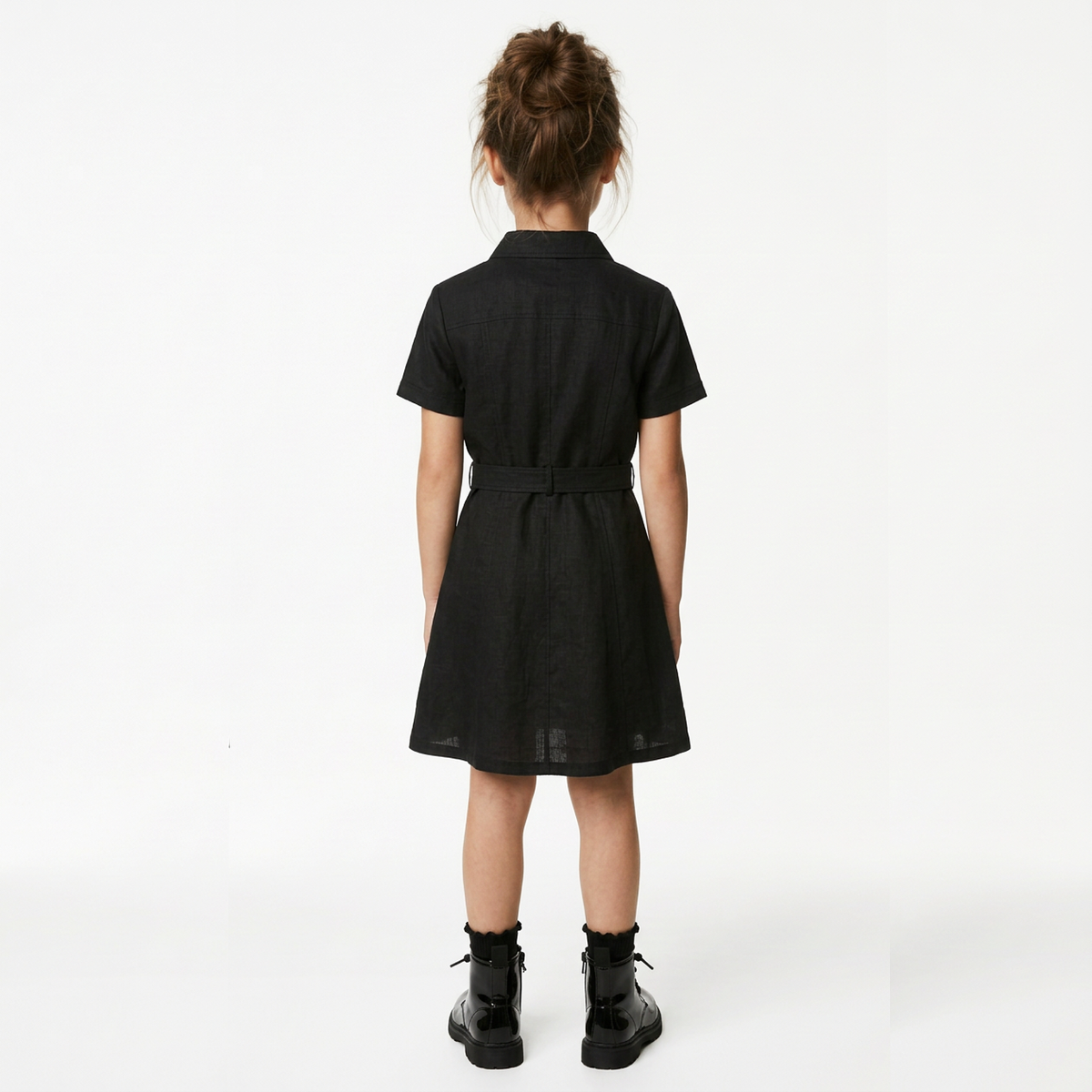 A child model shown from the back wearing a short-sleeved black collared dress with a matching tie belt and black ankle boots, photographed against a clean white studio background.