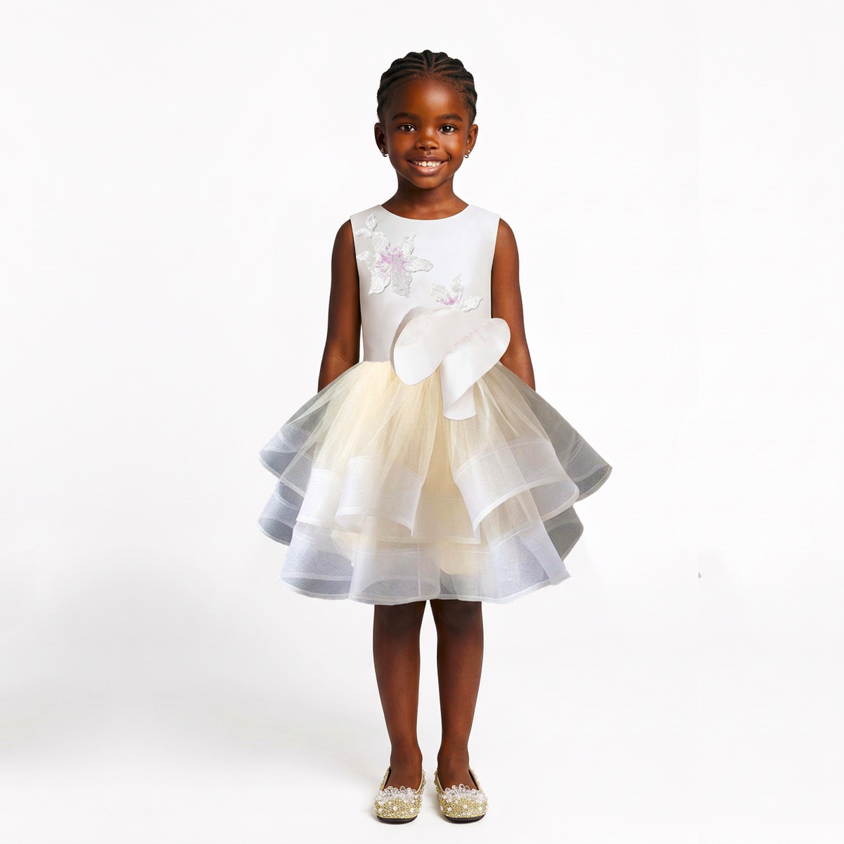 A young girl stands against a plain white studio background wearing a sleeveless white dress with a layered tulle skirt in white and pale yellow, a large bow at the waist, and floral embroidery near the neckline. She is also wearing sparkly flat shoes.