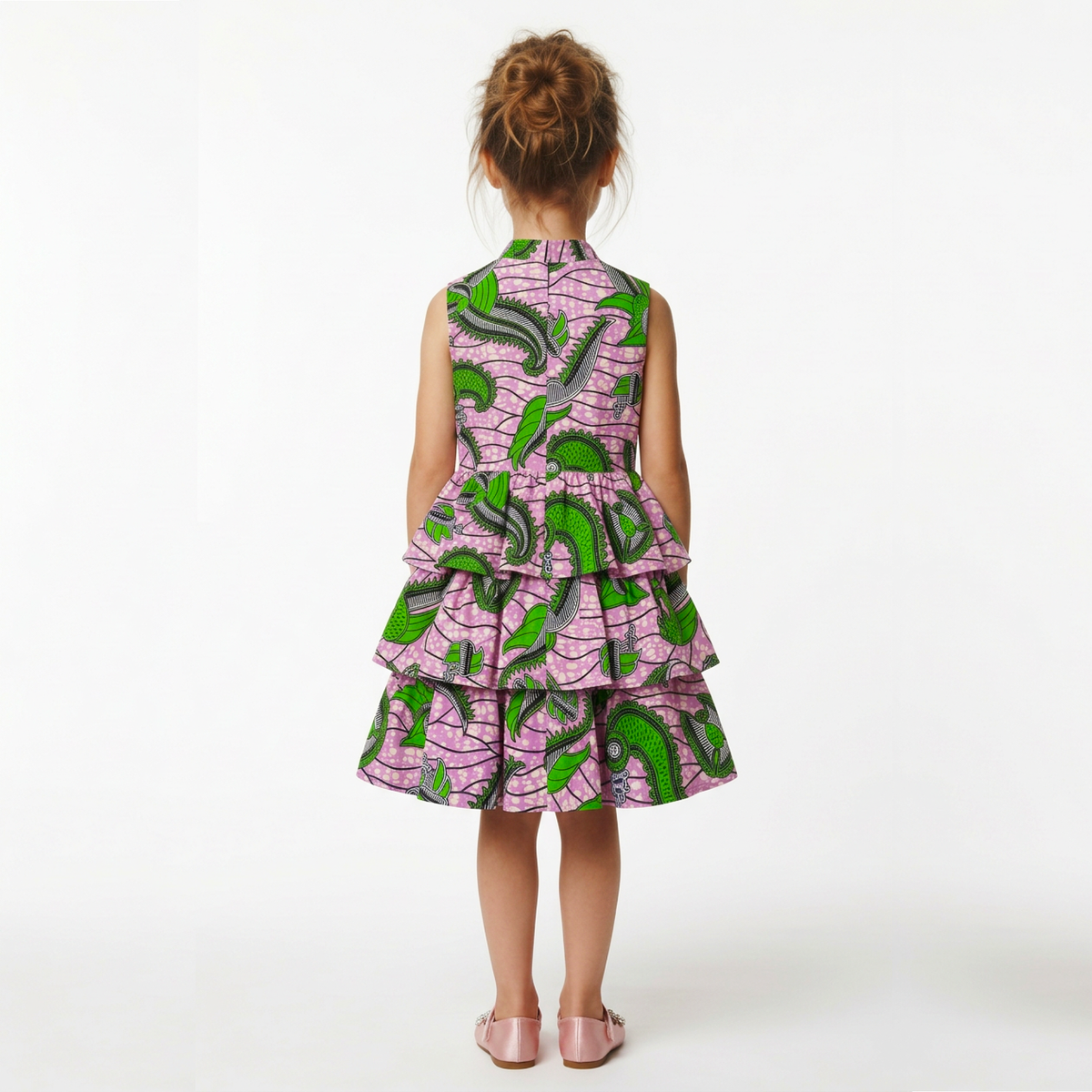A young girl viewed from behind wearing a sleeveless tiered dress with a pink and green abstract pattern, standing against a plain white studio background.