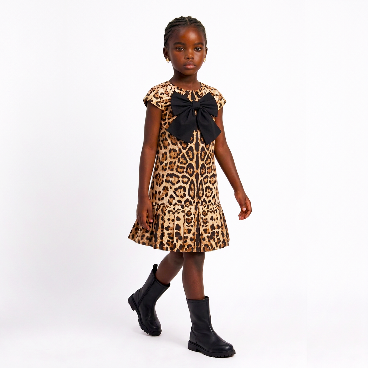 A young girl modeling a leopard print dress with a large black bow at the neckline and black ankle boots, standing against a plain white studio background.