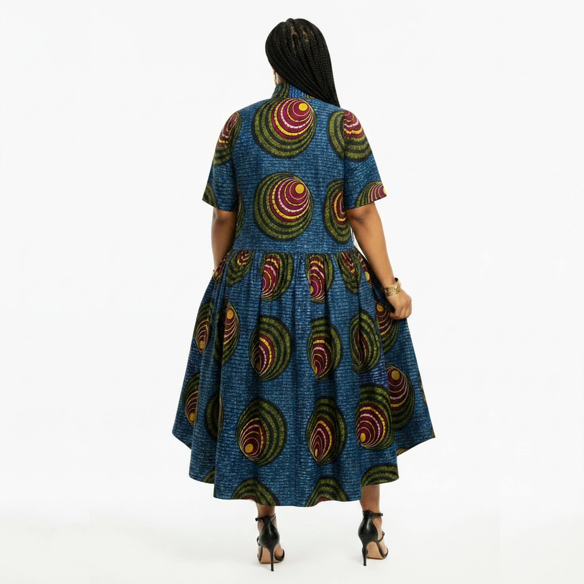 A woman viewed from the back wearing a short-sleeve midi dress with a blue base and circular multicolor African wax print, paired with black high-heeled sandals against a plain white background.