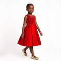 A young girl wearing a bright red sleeveless dress with a beaded floral bodice and a flared skirt, paired with gold flat shoes, standing against a plain white studio background.