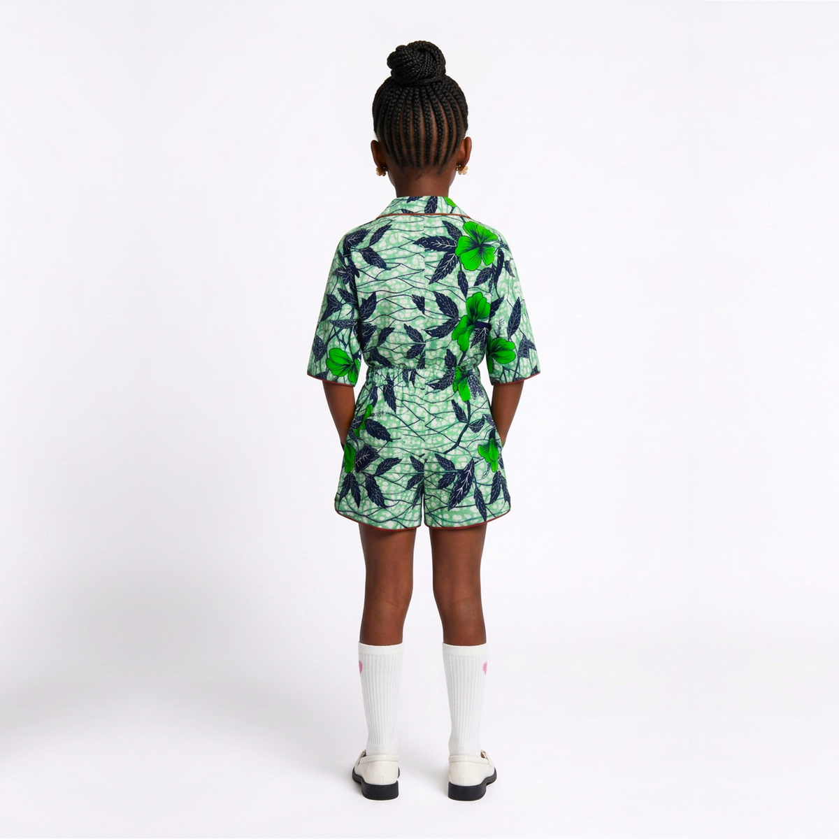 A child model viewed from the back wearing a short-sleeve collared romper with a green and navy tropical leaf pattern, paired with white knee-high socks and white shoes, against a plain white studio background.