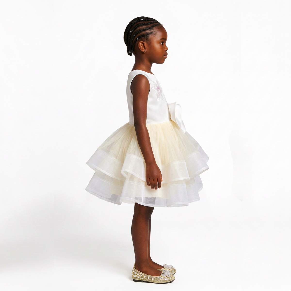 A young girl in profile wearing a sleeveless white and cream tulle dress with a tiered skirt and matching bow, standing against a plain white studio background.