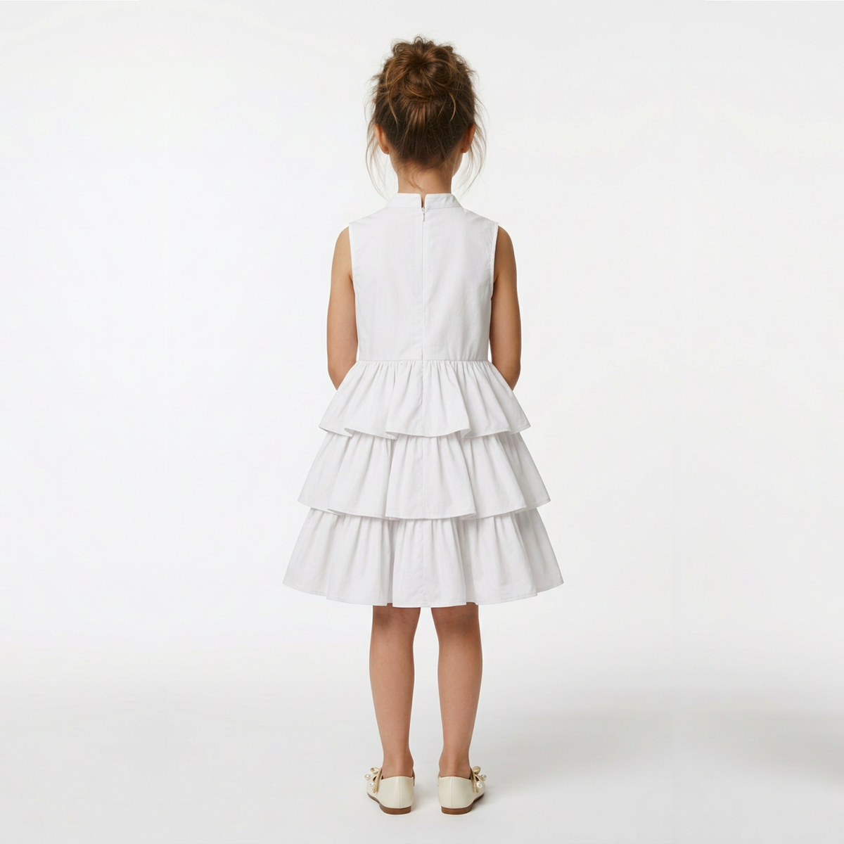 A young girl viewed from behind wearing a sleeveless white tiered dress and cream-colored shoes, standing against a plain white studio background.