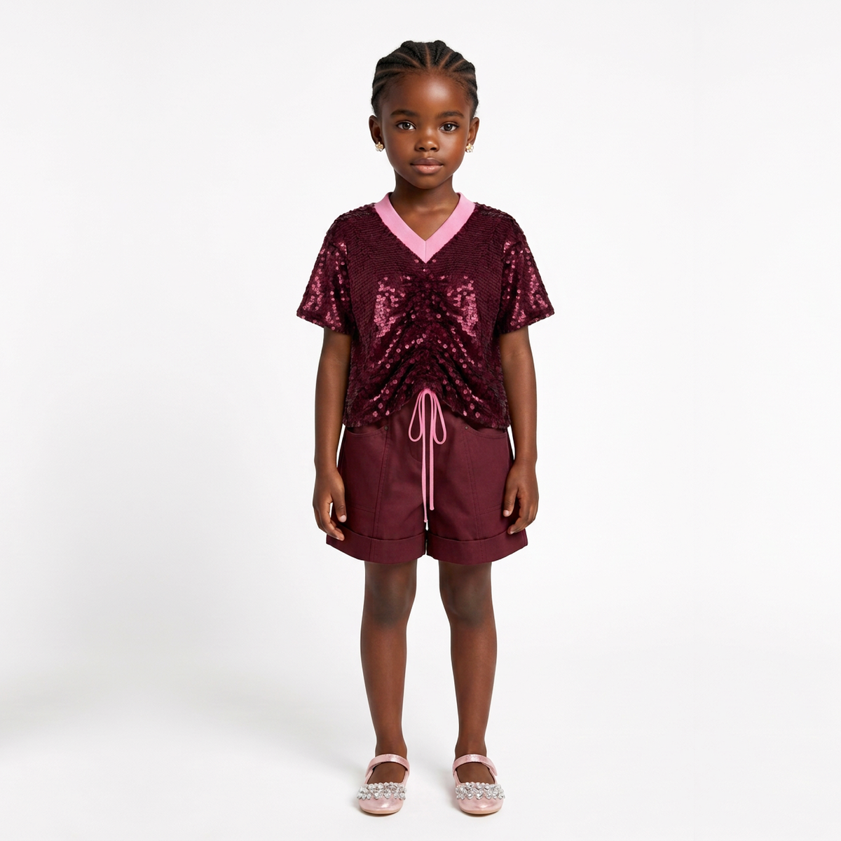 A young girl stands against a plain white studio background wearing a two-piece outfit consisting of a short-sleeve V-neck top covered in dark burgundy sequins with a light pink inner collar and matching pink drawstring, paired with matching burgundy shorts.