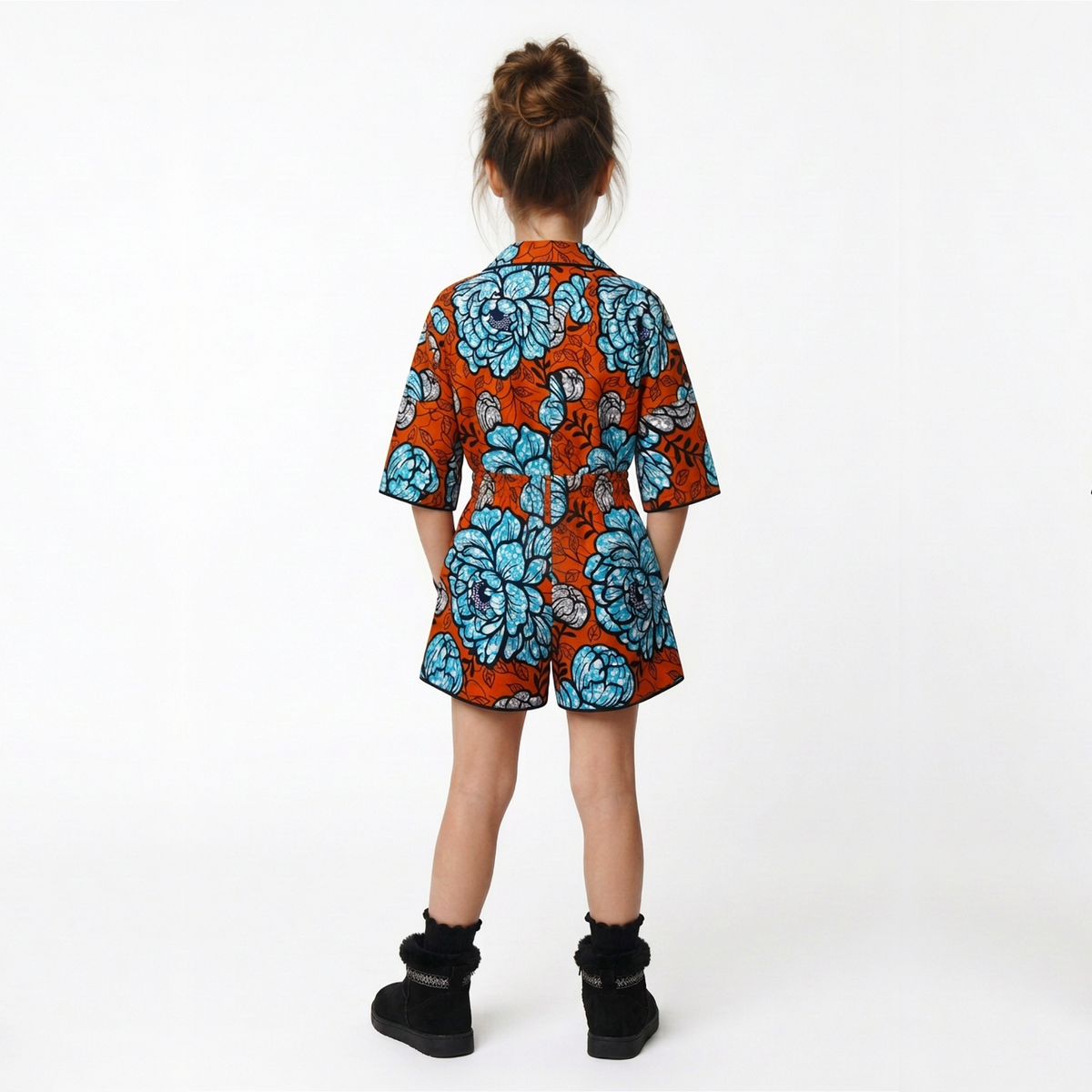 A child viewed from behind wearing a short-sleeve button-up shirt and matching shorts set in a vibrant orange fabric with a large blue floral print, paired with black ankle boots, against a plain white studio background.