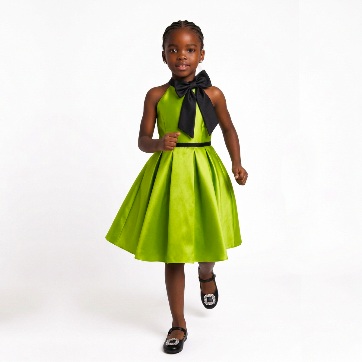 A young girl wearing a bright lime green halter dress with a large black bow at the neckline and a black waistband, paired with black flat shoes, posing against a white studio background.