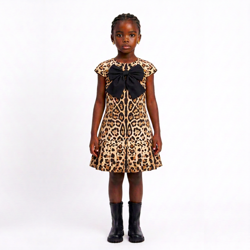 A young girl modeling a leopard print dress with a large black bow at the neckline, paired with black boots, against a plain white studio background.