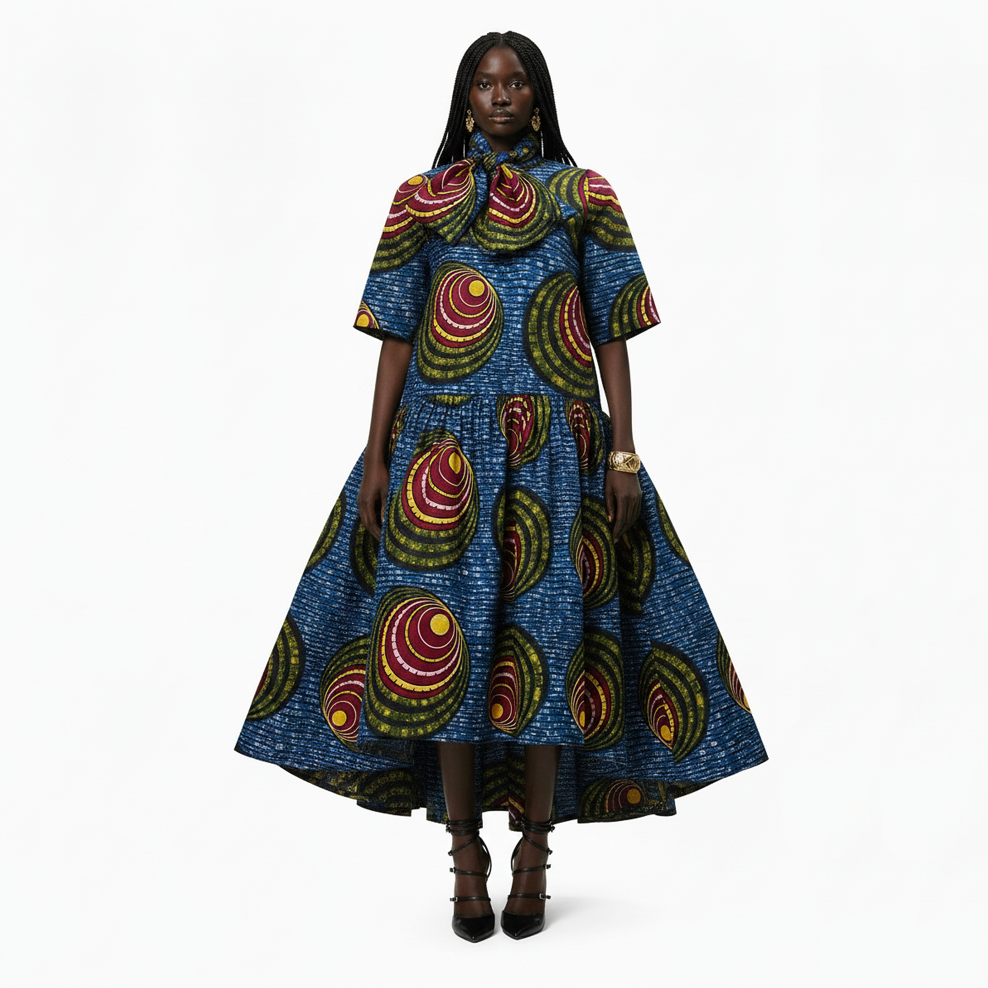A model stands against a plain white studio background wearing a high-waisted, short-sleeved midi dress with a vibrant blue base and a bold, circular African wax print pattern in red, yellow, and green.