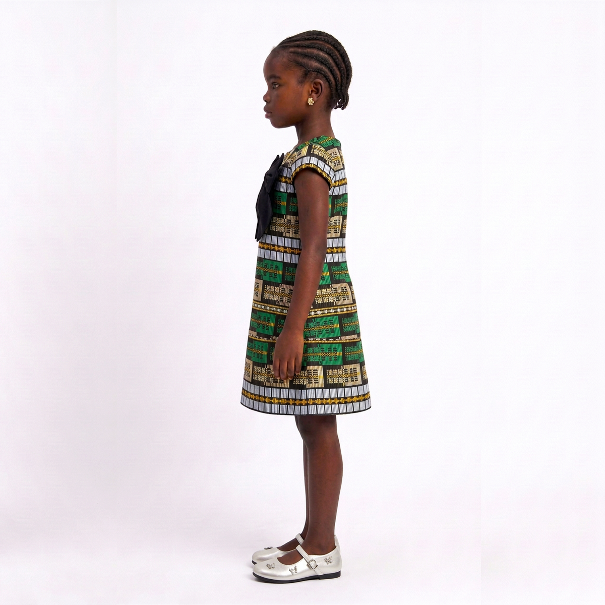 A young girl with braided hair stands in profile wearing a sleeveless A-line dress with a green, beige, and white geometric pattern and a black bow at the neckline, paired with white Mary Jane shoes, against a plain white studio background.