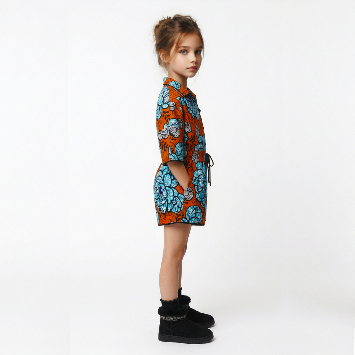 A young girl stands in profile wearing a short-sleeve collared shirt dress with a vibrant orange and blue floral pattern, paired with black ankle boots, against a plain white studio background.