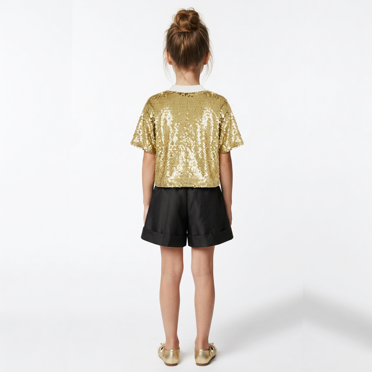 A young girl viewed from behind wearing a gold sequined short-sleeve crop top, black shorts, and gold flats, standing against a plain white studio background.