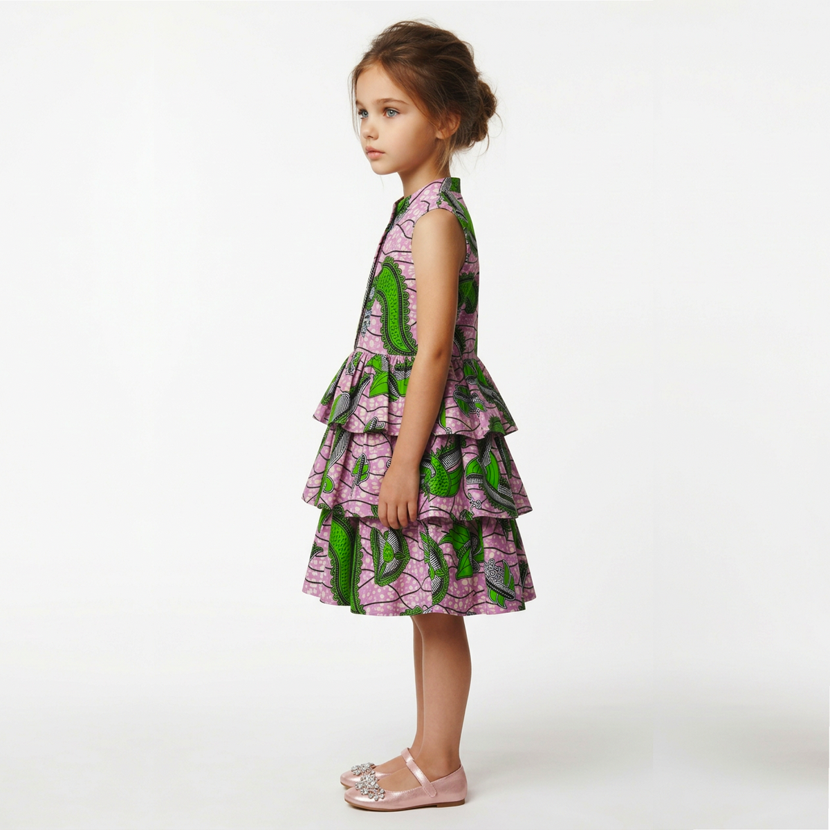 A young girl with her hair in a bun stands in profile wearing a sleeveless tiered dress with a pink and green leaf print, paired with light pink Mary Jane shoes, against a plain white studio background.