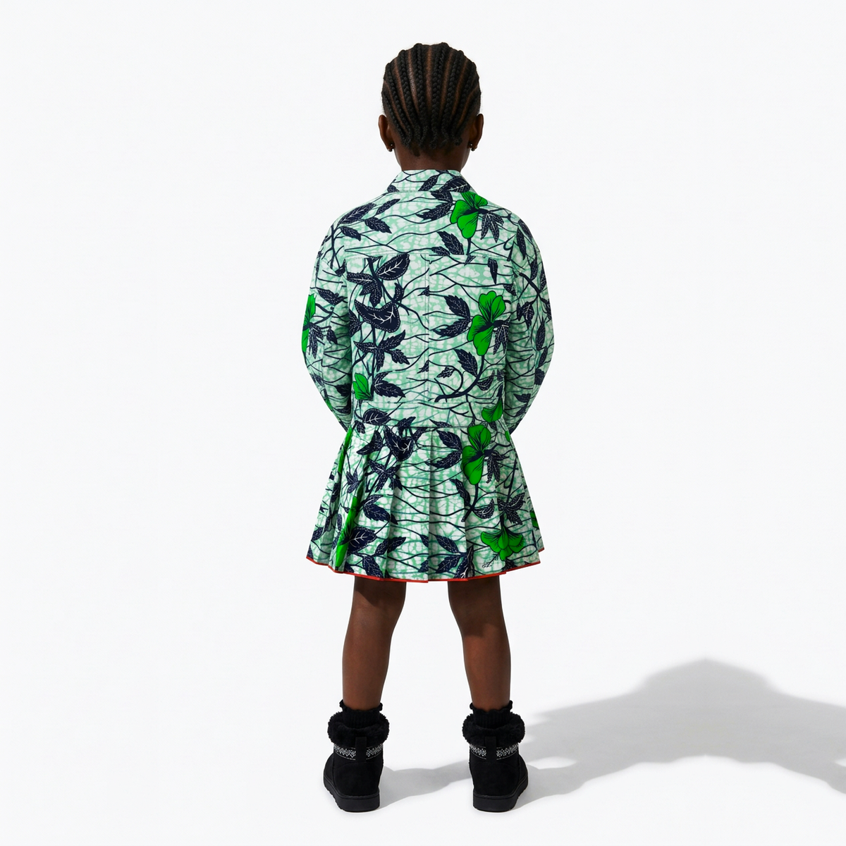 A child model shown from the back wearing a short green and black patterned coat or dress with a light mint background, paired with black boots, against a plain white studio background.