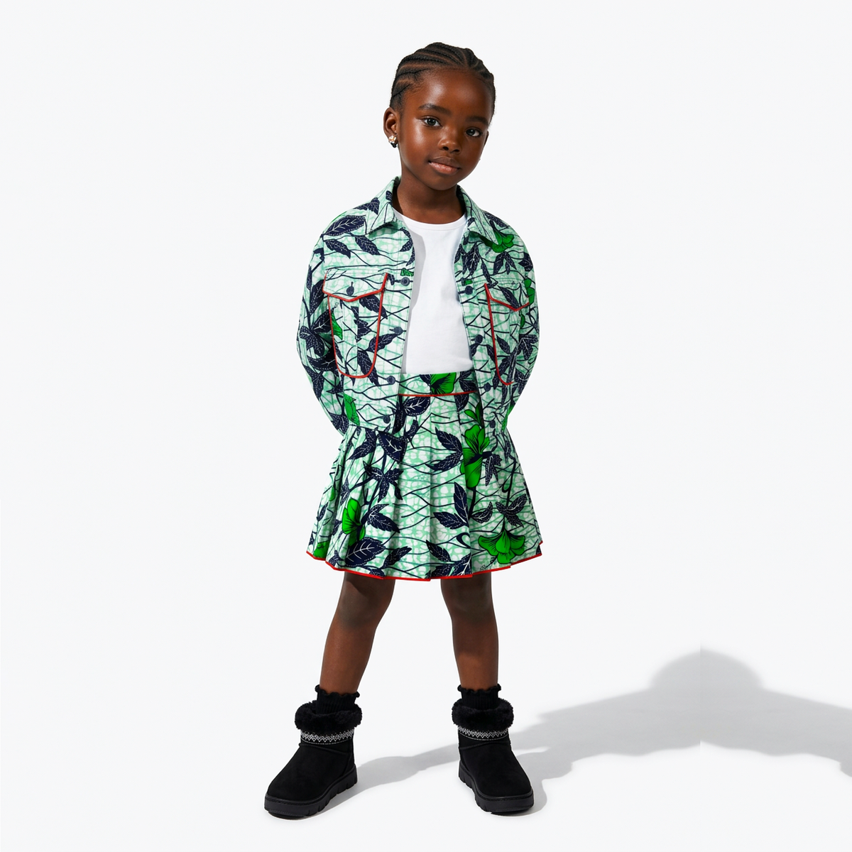 A young girl stands against a light gray studio background wearing a short printed jacket and matching skirt set. The outfit has a white base with a green and black leaf pattern and small red accents, paired with a white top and black ankle boots.