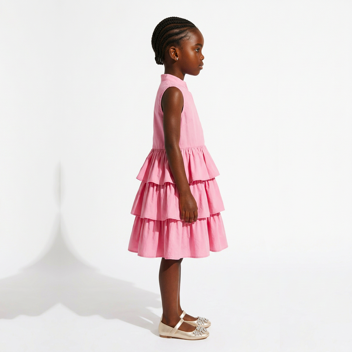 A young girl with braided hair stands in profile wearing a sleeveless tiered pink dress and beige flat shoes against a clean white studio background.