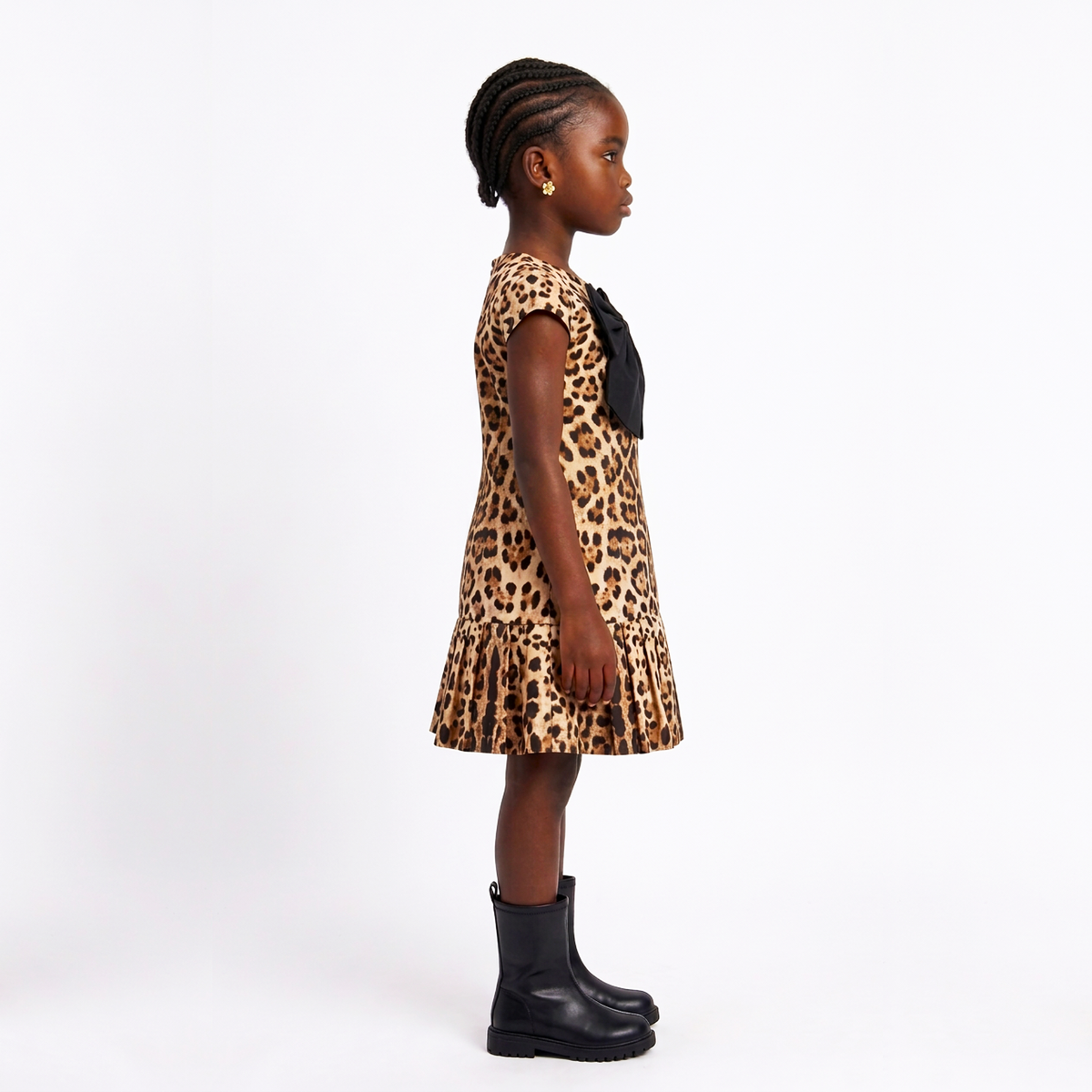 A young girl standing in profile wearing a leopard-print dress with a black bow at the neckline and black ankle boots, photographed against a plain white studio background.
