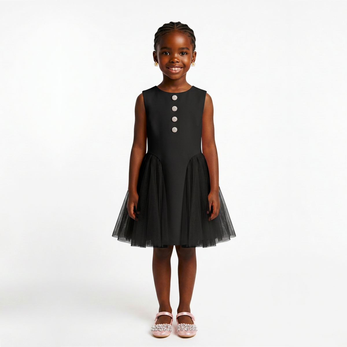 A young girl modeling a sleeveless black dress with a tulle skirt and three silver buttons on the chest, standing against a plain white studio background.
