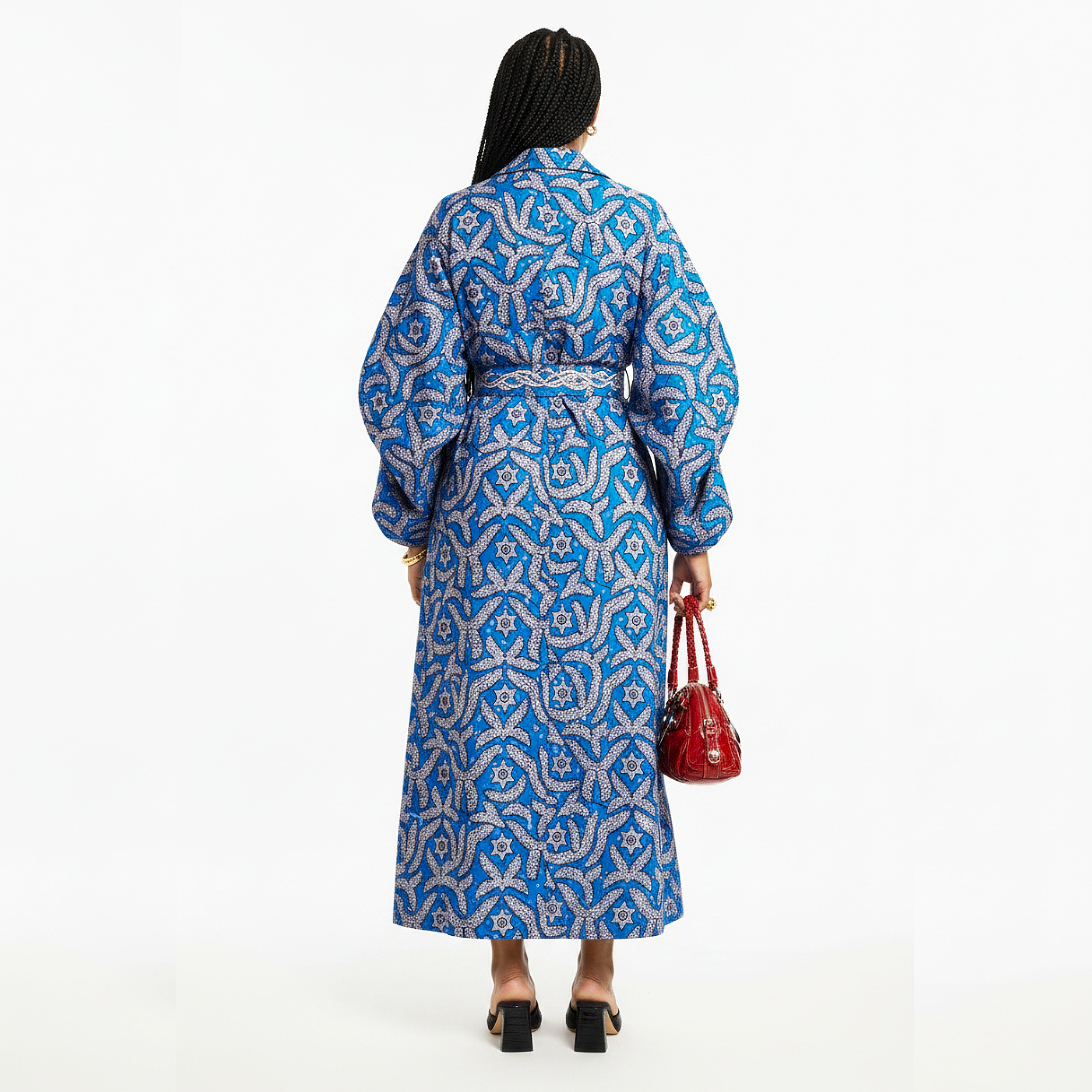 A woman shown from the back wearing a long-sleeved blue and white patterned maxi dress with a matching waist tie, holding a small red handbag against a plain white background.