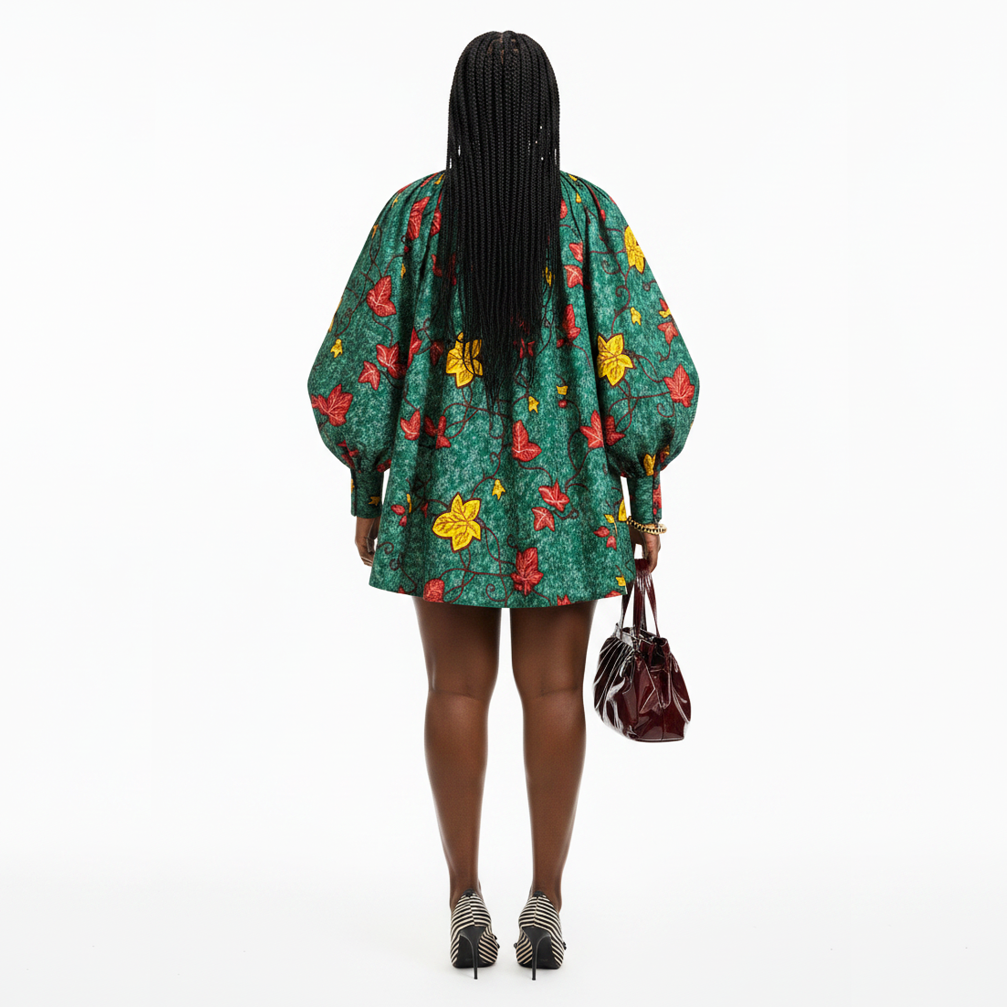 A woman is shown from behind wearing a green short puff-sleeve dress with a red and yellow floral pattern, holding a dark brown handbag against a solid white background.