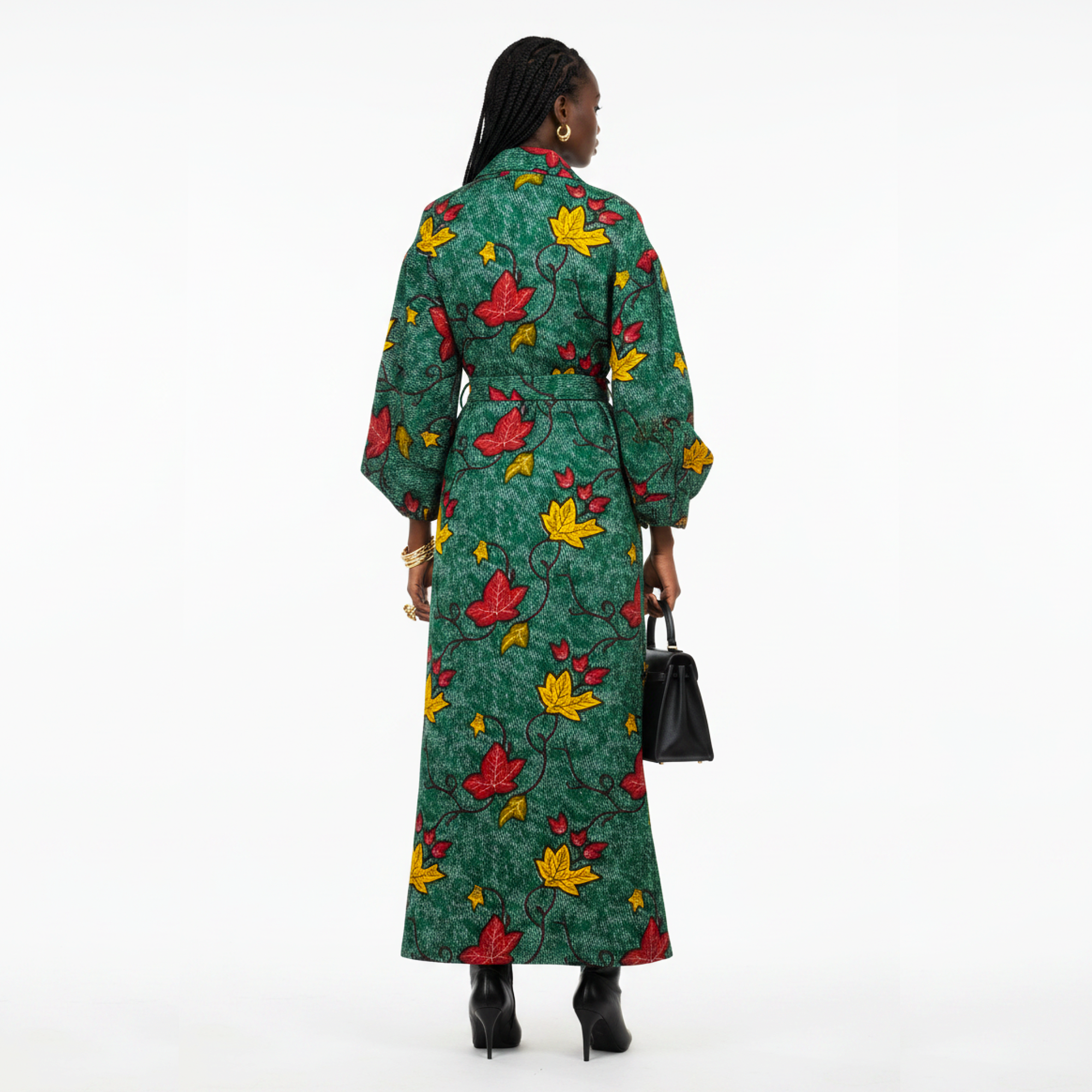 A woman viewed from the back wearing a long green dress with a red and yellow floral print, holding a small black handbag against a plain white studio background.