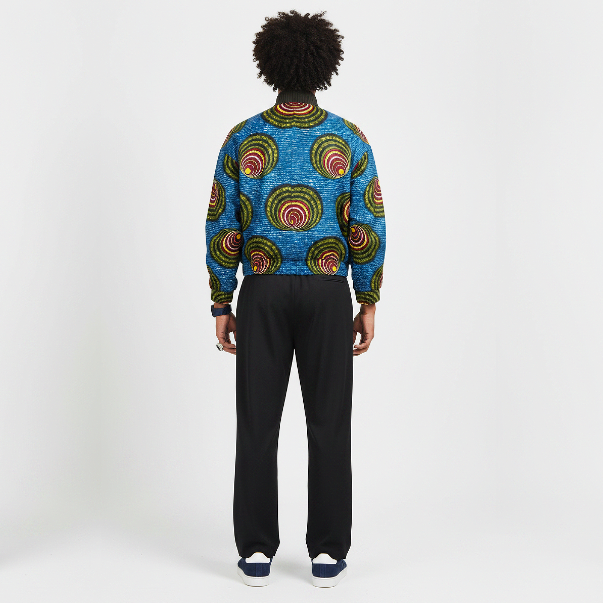 A model viewed from the back wearing a vibrant blue long-sleeve knit sweater with a bold circular African wax print, paired with black trousers and navy sneakers, against a plain white studio background.