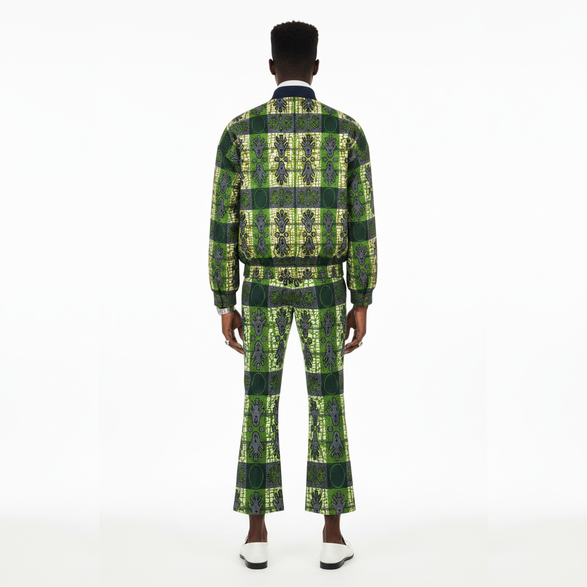A man viewed from the back wearing a matching green and yellow patterned two-piece suit with a long-sleeve jacket and flared trousers, paired with white loafers, against a plain white studio background.
