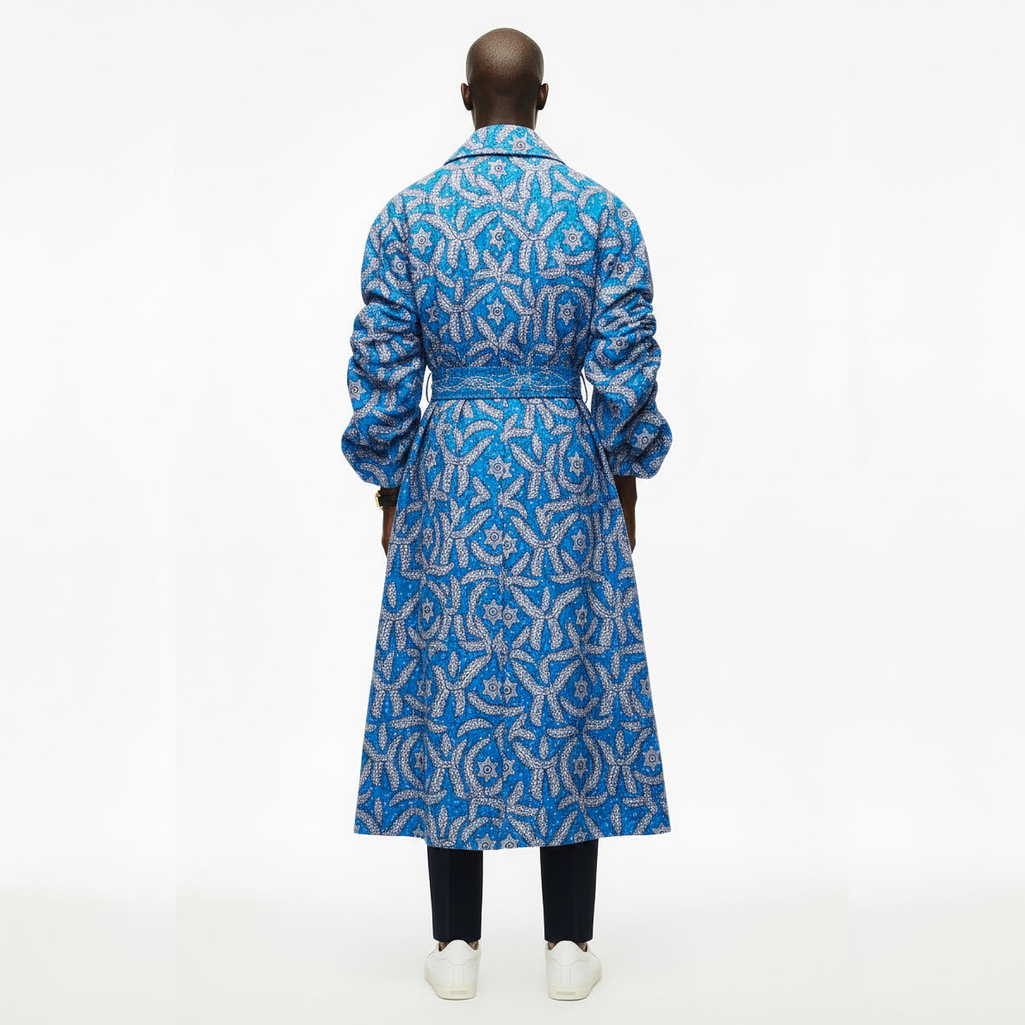 A model viewed from the back wearing a long blue and silver patterned coat, paired with black trousers and white sneakers, against a plain white studio background.